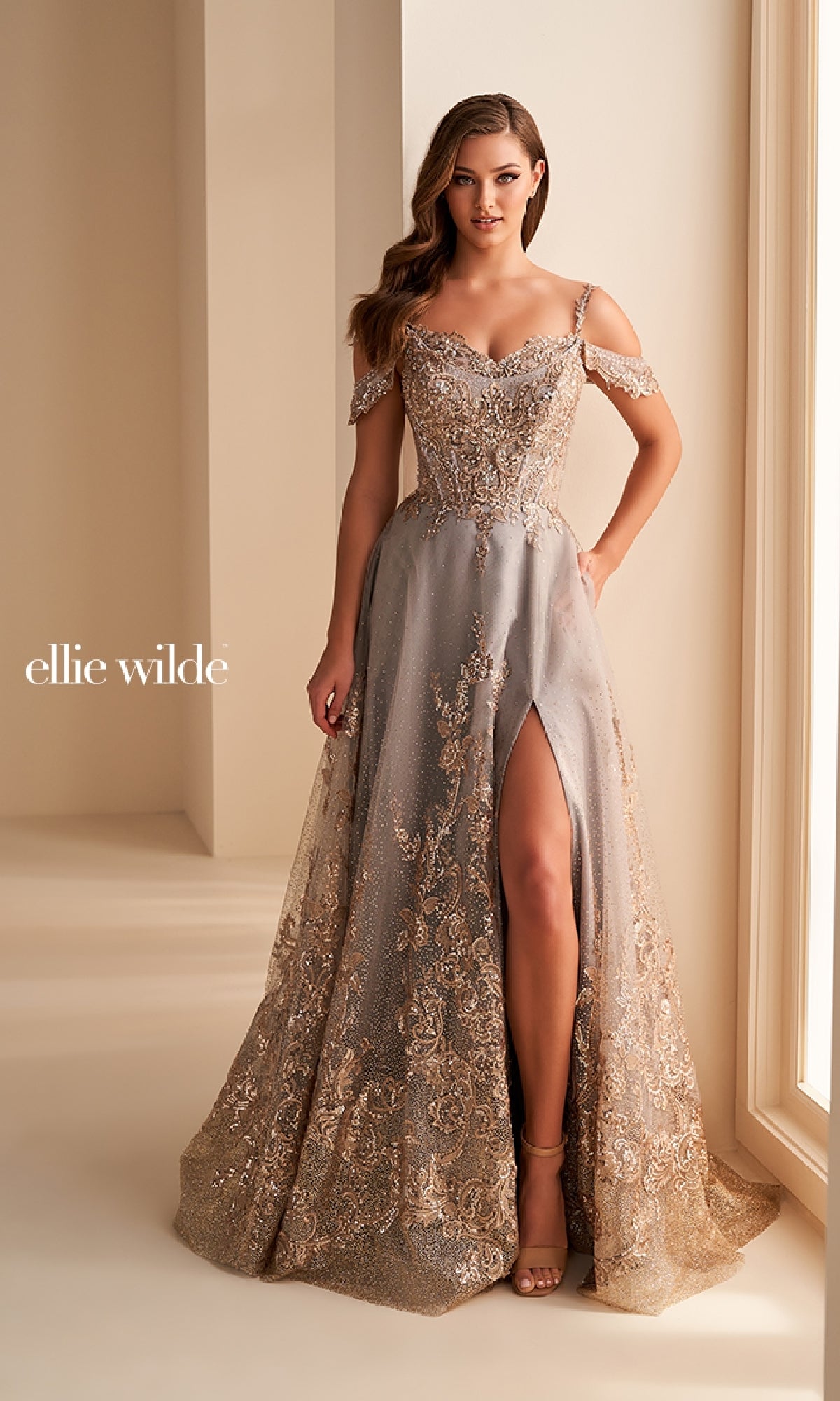 A woman stands indoors near large windows wearing the Ellie Wilde EW36220 Long Prom Dress, an off-the-shoulder gown with silver and gold embellishments, a thigh-high slit, glitter tulle, and detailed embroidery.